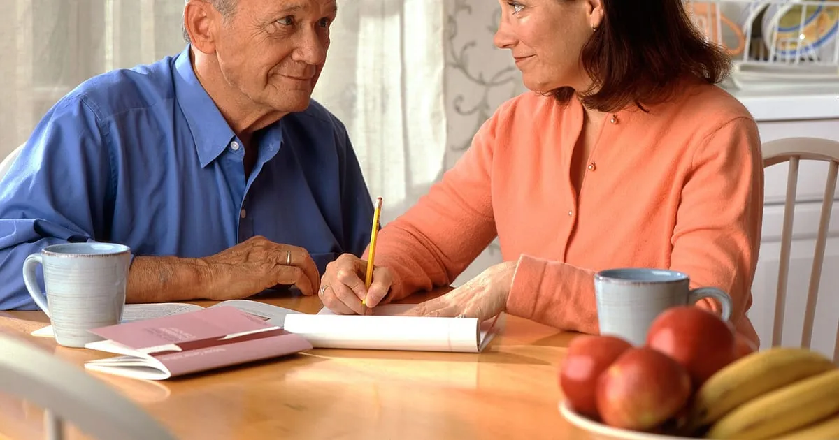 An older man and a woman sitting at a kitchen table with a notepad and paperwork, going over household costs